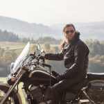 Handsome bearded motorcyclist with long hair in black leather jacket and sunglasses sitting on cruiser motorcycle, on blurred background of green peaceful rural landscape and light foggy sky.