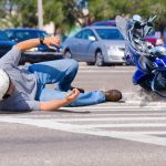 Man in helmet fallen on crosswalk beside a tipped-over blue motorcycle.